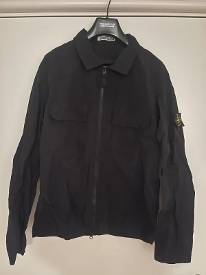 Svart overshirt från Stone Island - Svart overshirt från Stone Island med klassisk krage och dragkedja framtill. Jackan har två stora bröstfickor. Materialet känns lätt och har en snygg struktur, perfekt för lager-på-lager. Hör av dig för frågor eller funderingar!