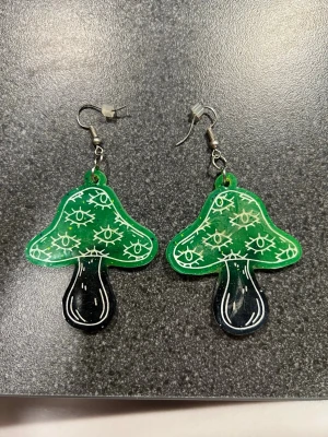 Grön svarta svamp örhängen med ögon - Unik örhängen i form av en grön svart svamp med vita ögon över hela ovandelen. Örhängena har en cool, konstnärlig vibe. Svampens fot är svart med vita detaljer. Perfekt för dig som gillar statement-plagg och vill sticka ut. De har fått ett litet konstigt lager på sig, kolla noggrant på bilderna.