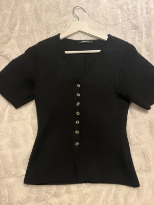 Svart v-ringad topp från Gina Tricot - Svart topp från Gina Tricot med v-ringning och korta ärmar. Toppen har en rad med silvriga knappar framtill och är figurnära i modellen. Materialet känns mjukt och stretchigt, perfekt för en clean och stilren look. Använd två gånger, som ny.
