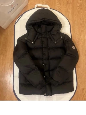Svart dunjacka från Moncler med huva - Svart dunjacka från Moncler med stor huva och klassisk logga på ärmen. Jackan har quiltad design, knappar och dragkedja framtill samt elastiska muddar vid ärmslut. Insidan är glansig och jackan har en loose passform som ger en cool vibe.