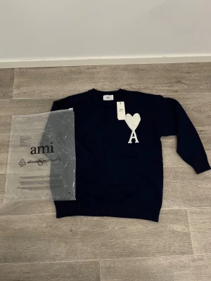 Mörkblå stickad tröja från Ami - Snygg mörkblå stickad tröja från Ami Alexandre Mattiussi med vit hjärtformad A-logga på bröstet. Tröjan har rund hals, ribbade muddar och är långärmad. Perfekt för dig som gillar stilrena detaljer och vill ha något som sticker ut lite extra.