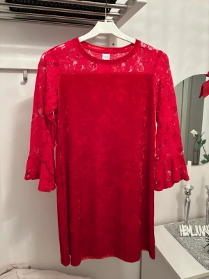 Röd spetsklänning med volangärm - Snygg röd klänning från Lindex med långa spetsärmar och volangdetaljer vid ärmslut. Klänningen har en rund halsringning och är tillverkad i ett mjukt, glansigt tyg med spets upptill och på ärmarna. Perfekt för dig som vill sticka ut med färg och detaljer.