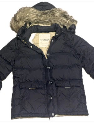 Svart pufferjacka från Ralph Lauren - Svart pufferjacka från Denim & Supply Ralph Lauren med beige foder och stor huva med fejkpäls. Jackan har två stora fickor framtill, dragkedja och knappar. Perfekt för kalla dagar med sin quiltade design och fluffiga känsla.