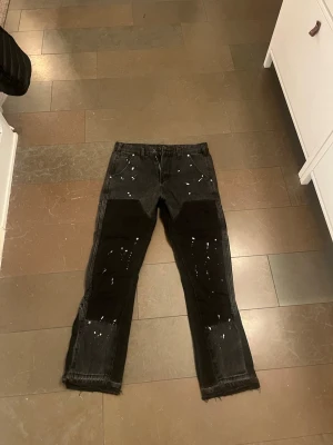Svarta jeans med färgstänk från Five Leafs - Unika svarta jeans från Five Leafs med vita färgstänk och råa, fransiga benslut. Byxorna har raka ben, klassiska fickor fram och bak samt extra fickor på sidorna. Materialet är robust jeans och designen har en cool, street-inspirerad vibe. Perfekt för dig som vill ta din stil till nästa nivå med unika flared jeans! Dem är helt oanvända med prislapp.