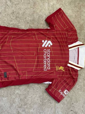 Ingen sko på bild - Bilderna visar en röd fotbollströja från Liverpool FC med gula och vita detaljer, samt sponsortryck. Ingen sko finns på bilderna.