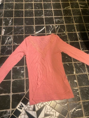 Rosa långärmad t-shirt med spets - Säljer en rosa långärmad t-shirt med snygg v-ringning och spetsdetalj runt halsen. T-shirten är gjord i mjuk bomull och har en feminin, normal passform. Perfekt för dig som gillar enkla plagg med en liten twist.