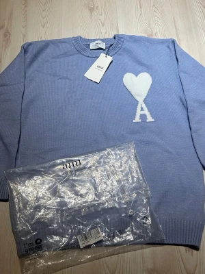 Ljusblå stickad tröja från Ami Paris- Storlek: S - Säljer en ljusblå stickad tröja från Ami Alexandre Mattiussi med ett stort vitt hjärta och bokstaven A på bröstet. Tröjan har rund hals, ribbade muddar och lång ärm. Perfekt för dig som gillar stilrena detaljer och snygg design.