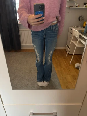 Blå bootcut jeans med slitningar - Säljer ett par blå jeans med bootcut och snygga slitningar framtill. Jeansen har låg midja och detaljerade bakfickor med knappar och dekorativa sömmar. Materialet är klassisk jeans och passformen är normal. Perfekt för dig som gillar en avslappnad och trendig stil. 🙂💞🫰🏼