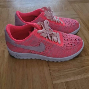 Nike Air Force 1 Flyknit rosa sneakers - Nike Air Force 1 Flyknit sneakers i en fräsch rosa färg med ljusrosa detaljer och vit sula. Skorna har stickad ovandel med ventilerande hål, klassisk snörning och rund tå. Swoosh-loggan i mocka på sidan och bakparti i ljusrosa mocka. Perfekt för dig som gillar färgstarka sneakers.
