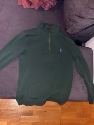 Mörkgrön half zip tröja Polo Ralph Lauren - Snygg mörkgrön långärmad tröja från Polo Ralph Lauren med half zip och liten lila logga på bröstet. Tröjan har en hög krage och är gjord i mjuk bomull, perfekt för kyliga dagar. Passar dig som gillar klassisk stil med en modern twist.