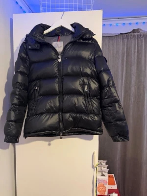 Svart pufferjacka från Moncler - Svart pufferjacka från Moncler med glansig finish och klassisk Moncler-logga på ärmen. Jackan har huva, två sidofickor med dragkedja och kraftig dragkedja framtill. Perfekt för kalla dagar och har en riktigt schysst streetstil. Är dags för nåt nytt.
