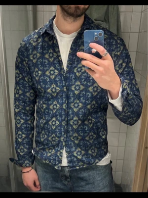 Blå mönstrad skjorta från Scotch & soda - Säljer en blå skjorta från scotch & soda med unikt blommigt mönster i gult och grått. Skjortan har klassisk krage, knappar framtill och långa ärmar. Materialet är mjukt och känns som bomull, perfekt för dig som vill sticka ut med en cool look.