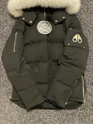 Svart Moose Knuckles dunjacka med päls - Svart pufferjacka från Moose Knuckles med fluffig vit päls på huvan, silverfärgade detaljer och logga på ärmen. Jackan har dragkedjor på sidorna, stora knappar och praktiska fickor framtill. Perfekt för kalla dagar och riktigt snygg stil och vattentät, pris kan diskuteras har använts få gånger väldigt bra skick