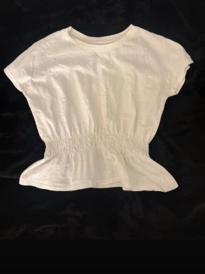 Vit topp med smock i midjan - Säljer en vit topp med pärlor och smockad midja som ger en snygg volangkant nedtill. Köpt på barnavdelningen strl 134-140, passar mig som har s/xs, cirka 165cm.