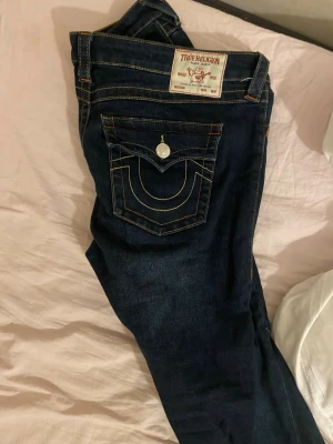 Mörkblå True Religion bootcut jeans - Snygga mörkblå jeans från True Religion med klassiska kontrastsömmar och ikoniska ficklock med silverfärgade knappar. Modellen har bootcut-ben och markerade bakfickor med broderad logga. Perfekt för dig som gillar statement-jeans med coola detaljer.