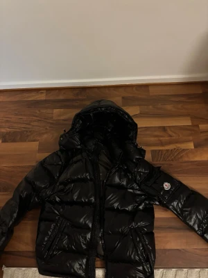 Moncler maya - Säljer en svart pufferjacka från Moncler med glansig finish och huva. Jackan har dragkedja framtill, två sidofickor med dragkedja och Moncler-logga på ärmen. Perfekt för kalla dagar och har en riktigt snygg streetstil.