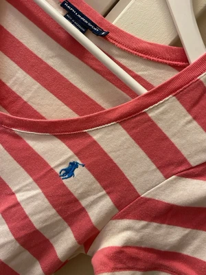 Randig t-shirt från Ralph Lauren - Säljer en fräsch t-shirt från Ralph Lauren Sport med breda rosa och vita ränder, v-ringad hals och korta ärmar. T-shirten har en blå broderad logga på bröstet och är gjord i mjuk bomull. Perfekt för dig som gillar klassisk stil med en twist.