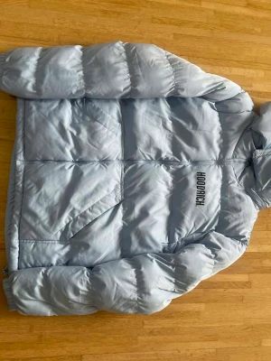 Ljusblå pufferjacka från Hoodrich - Säljer en ljusblå pufferjacka från Hoodrich med stor logga på ryggen och mindre logga på bröstet. Jackan har huva, dragkedja framtill och två snedställda fickor. Materialet är syntet och jackan är quiltad för extra värme. Perfekt för kalla dagar.
