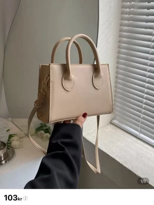 Beige axelväska i syntet - Stilren beige axelväska i syntetmaterial med två handtag och justerbar axelrem. Väskan har en rektangulär form och ett huvudfack med dragkedja. Perfekt storlek för det viktigaste och passar till många olika outfits.