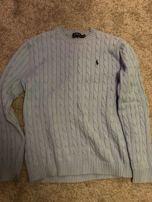Ljusblå stickad tröja från Polo Ralph Lauren - Säljer en ljusblå stickad tröja från Polo Ralph Lauren med klassiskt kabelstickat mönster och rund hals. Tröjan har långärmade ribbade muddar och en broderad logga på bröstet. Perfekt för dig som gillar stilrena och tidlösa plagg.
