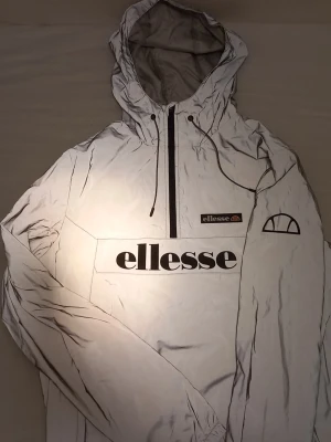 Vit vindjacka från Ellesse med huva - Säljer en vit vindjacka från Ellesse med stor logga framtill och snygga svarta detaljer. Jackan har huva med snörning, half zip och lång ärm. Materialet är 100% polyester och jackan har en sportig vibe som passar perfekt till streetwear.
