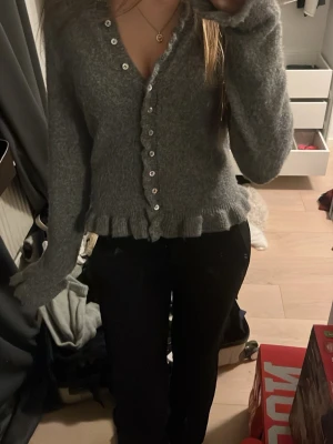 Grå kofta med volangkant - Säljer en mysig grå kofta med fluffig känsla och volangkant nedtill. Koftan har långa ärmar och knappar hela vägen framtill. Säljer eftersom den inte kommer till användning