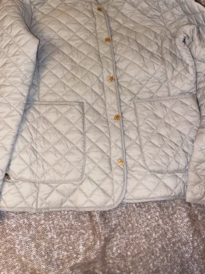 Beige quiltad jacka från Marine Classic - Quiltad beige jacka från Marine Classic med snygga guldfärgade knappar och stora fickor framtill. Jackan har ett klassiskt snitt och är perfekt för dig som gillar stilren och enkel design. Passar dig som vill ha en jacka med lite extra detaljer.