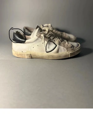 Vita sneakers med svart detalj - Säljer ett par vita sneakers med svart detalj på hälen och snörning framtill. Skorna har en låg profil och platt sula, samt en unik sydd detalj på sidan. Materialet är en mix av mocka och skinn vilket ger en schysst kontrast.
