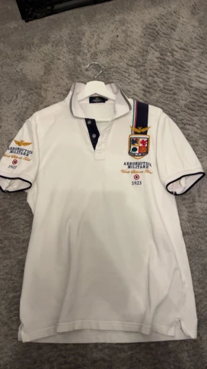 Vit Aeronautica Militare piké - Vit pikétröja från Aeronautica Militare med broderade detaljer och emblem på bröstet och ärmen. Tröjan har korta ärmar, knappslå framtill och marinblå kontrastdetaljer vid krage och ärmslut. Perfekt för dig som gillar sportig och stilren look. Passar S-M