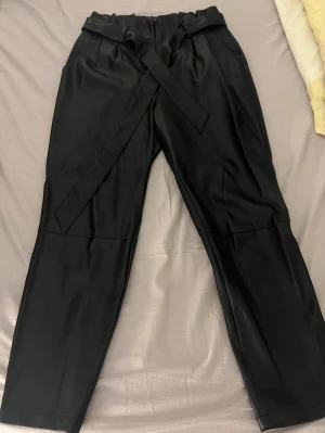 Svarta byxor i fuskskinn från Amisu/ NewYorker - Säljer ett par svarta byxor i fuskskinn från Amisu, storlek 40. Byxorna har hög midja, avsmalnande ben och ett brett bälte i midjan som knyts framtill. Dragkedja till gylf. Snyggt glansigt material och stilren look som passar till många outfits.