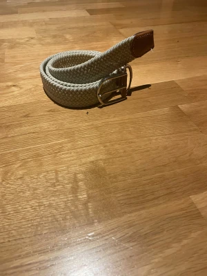 Beige flätad bälte med läderdetalj - Snyggt beige flätat bälte med silverfärgat spänne och en detalj i brunt läder vid änden. Perfekt för att ge outfiten lite extra stil och passar till både jeans och chinos. Bältet har en mjuk och flexibel känsla tack vare det stickade materialet.
