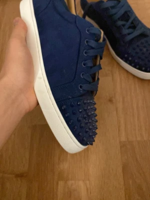 Mörkblå Christian Louboutin sneakers - Säljer ett par mörkblå sneakers från Christian Louboutin med klassisk röd sula och coola nitar på tån. Skorna har snörning, rund tå och är gjorda i mocka med vit platt sula. Perfekt för dig som vill sticka ut med exklusiv stil.