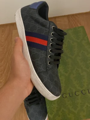 Gucci sneakers med blå och röd detalj - Säljer ett par svarta Gucci sneakers med diskret GG-mönster, blå och röd rand på sidan samt metallic-detaljer i blått och rött på hälen. Skorna har snörning och vit platt sula. Kommer med originalkartong. Perfekt för dig som gillar lyxiga streetwear-vibes.