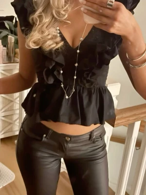 Svart blus med volanger och v-ringning - Säljer en svart populär blus med djup v-ringning och med volanger. Blusen har en smockad struktur längs axlarna och en peplum-liknande nederdel. Dragkedja i ryggen för enkel påklädning. 