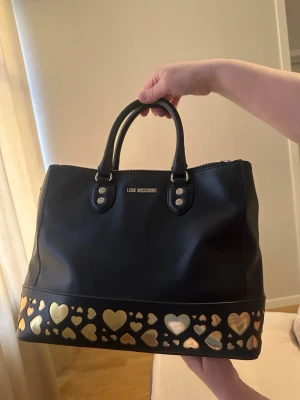 Svart handväska från Love Moschino - Säljer en svart handväska från Love Moschino med två stadiga handtag och dragkedja upptill. Väskan har en dekorativ kant med guldiga hjärtan längst ner som ger en unik och cool vibe. Materialet är skinn och detaljerna är i metall. Perfekt för dig som gillar statement-accessoarer.