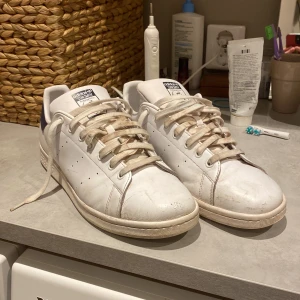 Adidas Stan Smith vita sneakers - Klassiska Adidas Stan Smith sneakers i vitt skinn med mörkblå detalj på hälen och perforerade ränder på sidorna. Skorna har snörning, platt sula och rund tå. Perfekta för dig som gillar en clean och tidlös look. Skick 8.5/10 lite smuts här å där följer tyvärr inte med någon box