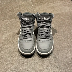 Grå adidas high top sneakers - Snygga grå high top sneakers från adidas med klassiska vita ränder på sidorna och tydlig adidas-logga på plös och häl. Skorna har snörning, vadderad krage och platt sula. Perfekta för dig som gillar sportig och streetinspirerad stil.