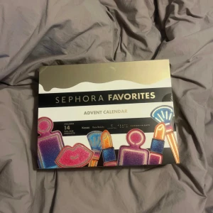 Sephora Favorites Adventskalender - Säljer en Sephora Favorites Adventskalender med 14 fullstora sminkprodukter från märken som Kosas och Rare Beauty. Kalendern har en lyxig design i guld, svart och färgglada illustrationer av smink, perfekt för dig som vill testa olika produkter.