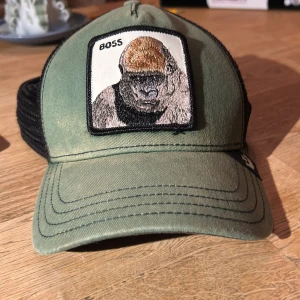 Grön Goorin Bros keps med gorilla-patch - Snygg grön keps från Goorin Bros med svart mesh baktill och justerbar snapback. Framtill sitter en broderad patch med en gorilla och texten 'BOSS'. Kepsen har cool streetstyle-vibe och är perfekt för dig som vill sticka ut. 