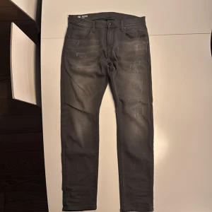 Svarta G-Star Revend jeans, W32 L30 - Snygga svarta G-Star Revend jeans med slitna detaljer och tvättad look. Modellen har fem fickor, smal passform och klassisk jeansdesign. Innerbensmått 72cm över midja 40,5cm.  Materialet är stretchigt för extra komfort och byxorna har diskreta slitningar framtill för en cool vibe.