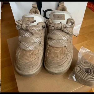 Beige sneakers från UGG med mönstrade snören - Snygga beige sneakers från UGG med breda, mönstrade snören i beige och vitt. Skorna har en mix av mocka och textilmaterial, rund tå och platt sula. Perfekta för dig som gillar en clean och trendig stil. Extra snören medföljer.
