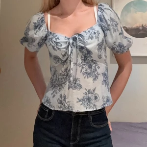 Blommig blus med puffärm  - Supersöt blus i ljusblått med mörkblått blommönster från vero moda. Toppen har puffiga korta ärmar, söt snörning framtill och fyrkantig ringning. Passformen är normal och toppen har en romantisk vibe. Köptes för 379 kr, aldrig använd!! 