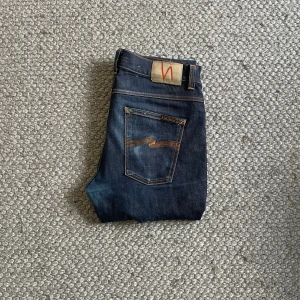 Mörkblå jeans från Nudie Jeans - Snygga mörkblå jeans från Nudie Jeans med klassisk orange söm och vågig detalj på bakfickorna. Modellen har raka ben och normal passform. Jeansen är tillverkade i slitstarkt bomullsjeansmaterial och har en klassisk femficksdesign.