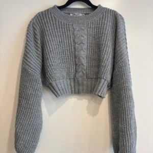 Grå croppad stickad tröja NA-KD - Snygg grå croppad stickad tröja från NA-KD med grov ribbad struktur och flätad detalj framtill. Tröjan har rund halsringning och långa ärmar, perfekt för att ge din outfit en chill vibe. Passar dig som gillar trendiga och bekväma plagg. Fin med skjorta under. 