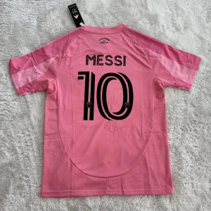 Inter Miami Messi #10 rosa matchtröja - Säljer en officiell Inter Miami fotbollströja från Adidas med Messi och nummer 10 på ryggen. Tröjan är rosa med svarta detaljer, har korta ärmar och randigt mönster framtill. Materialet är lätt och andas, perfekt för träning eller match. Klubbmärke och Adidas-logga på bröstet.