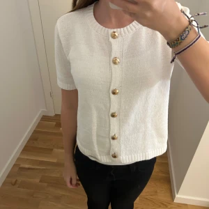 Vit stickad kofta från Sinsay - Vit stickad kofta från Sinsay i storlek S-passar XXS eller XS. Kortärmad med rund hals och snygga guldfärgade knappar framtill. Ribbad kant vid hals och nederdel ger en clean look. 