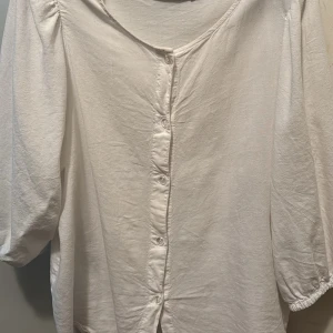 Vit blus med knappar framtill - Stilren vit blus med knappar längs hela framsidan och rundad halsringning. Blusen har trekvartsärm och är gjord i ett mjukt, tunt tyg som ger en avslappnad look. Perfekt att styla med jeans eller kjol för en enkel men snygg outfit.
