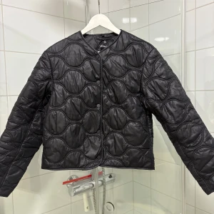 Jacka Mango - Svart quiltad pufferjacka från Mango med vågigt mönster i sömmarna. Jackan har rund halsringning, tryckknappar framtill och är tillverkad i glansig polyester. Perfekt för dig som vill ha en stilren och trendig look.