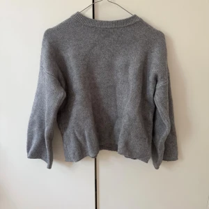Grå stickad tröja med rund hals - En enkel och stilren grå stickad tröja med rund halsringning och långa ärmar. Tröjan har en avslappnad passform och ribbade muddar vid halsen. Perfekt att slänga på sig när det är kyligt ute.