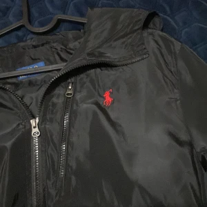 Svart vindjacka från Polo Ralph Lauren - Svart vindjacka från Polo Ralph Lauren med röd broderad logga på bröstet. Jackan har huva, dragkedja framtill och en bröstficka med dragkedja. Tillverkad i glansig polyester som ger en clean och sportig vibe.